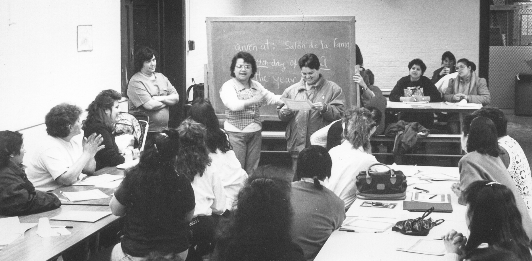 Una foto en blanco y negro de un grupo de mujeres en un salón de clases. Dos están parados cerca de la pizarra, una mujer habla y la otra sostiene un certificado.