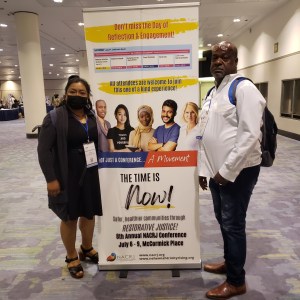 A Latina woman and African-American man stand in front of a sign for the NACRJ Conference in Chicago