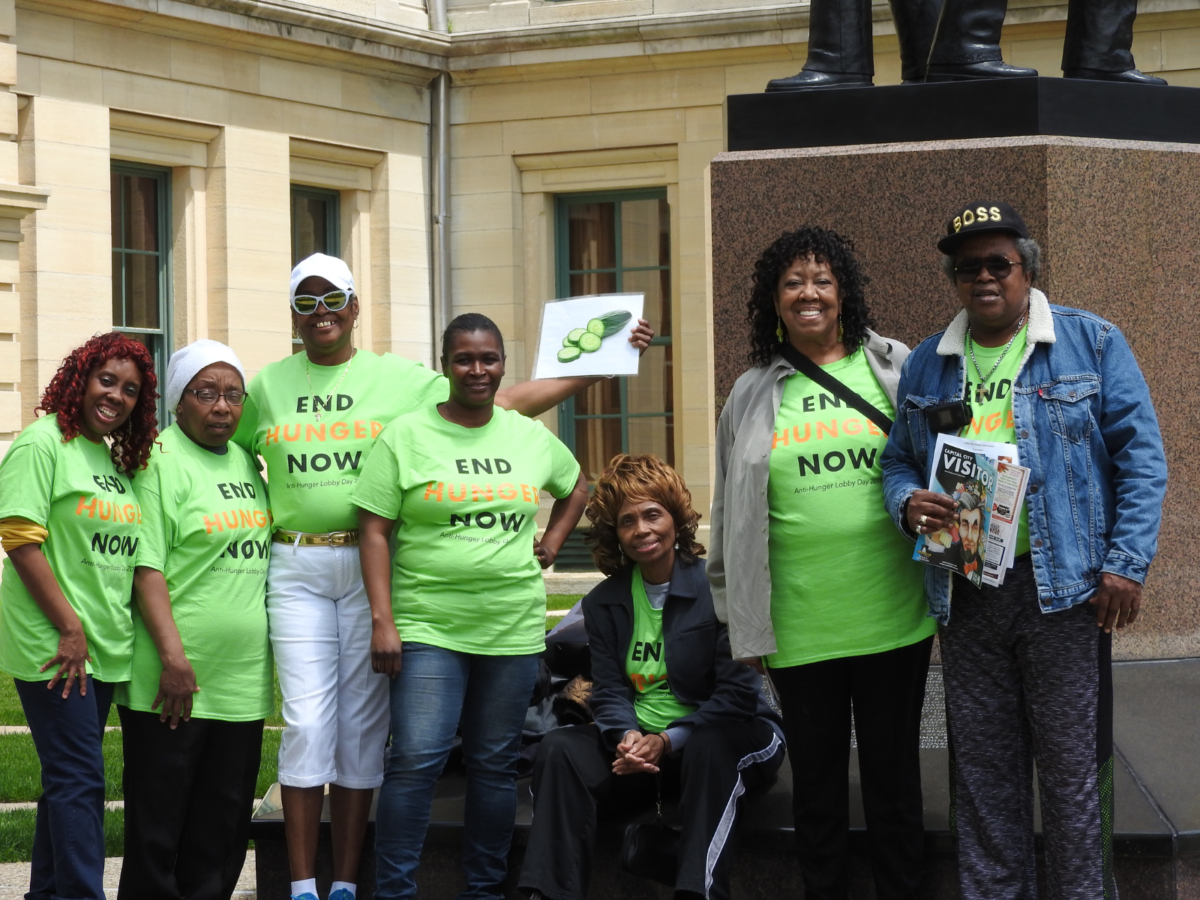 Un grupo de mujeres afroamericanas con camisas verdes que dicen End Hunger Now frente a un edificio del gobierno