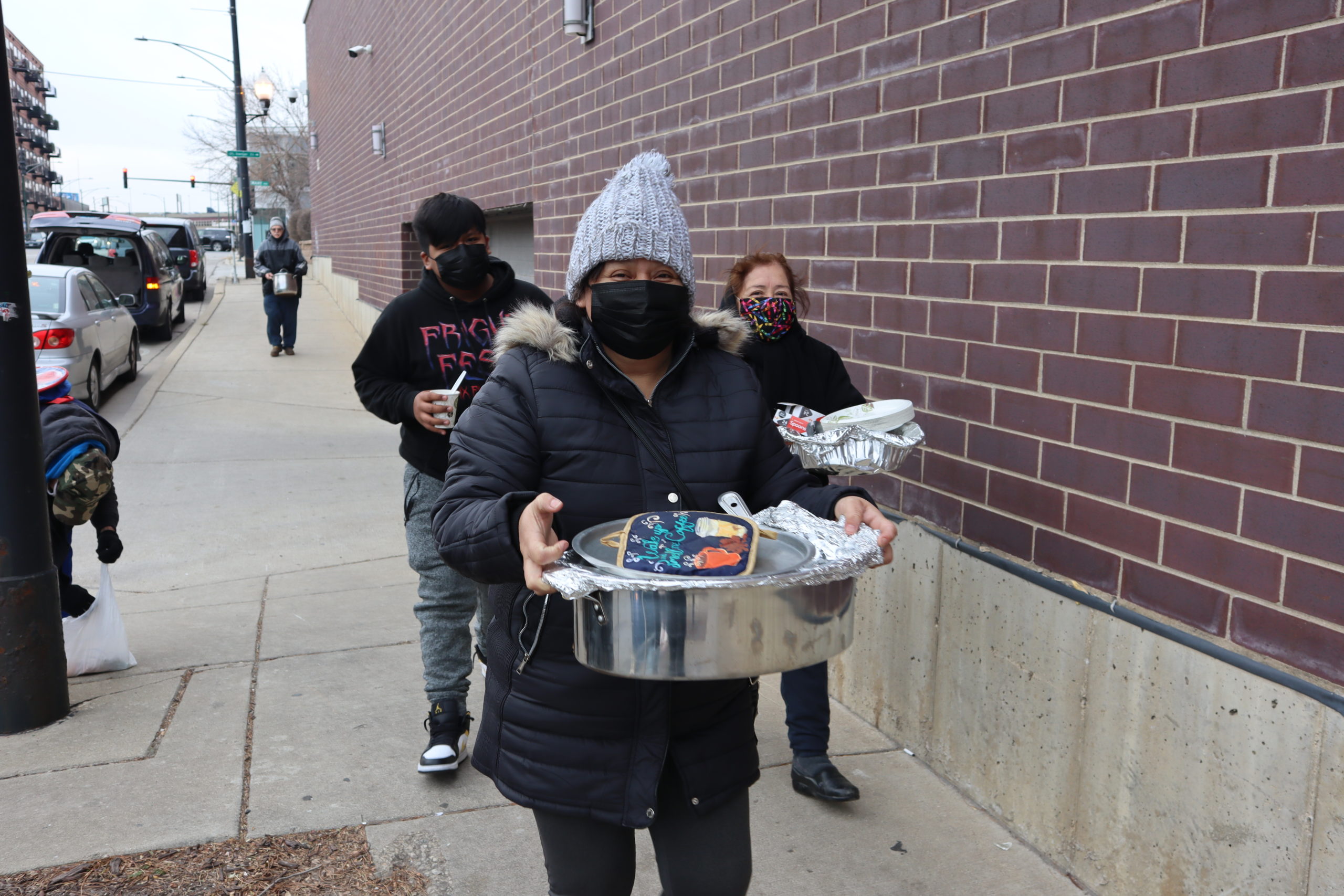 Three people are wearing masks and winter clothing; they're walking with pans of food