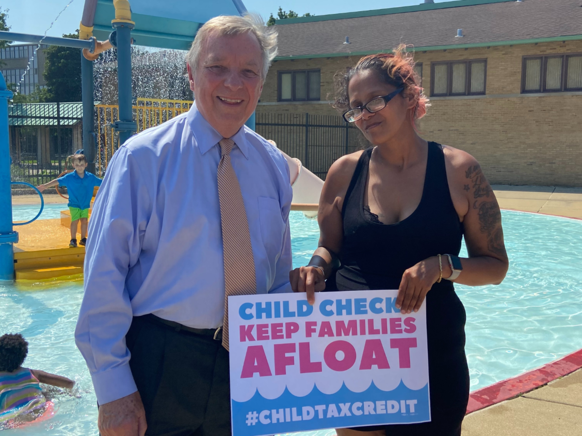 Una madre latina y una senadora estadounidense frente a una piscina. La mujer sostiene un cartel que dice Child Checks Keep Families Afloat.