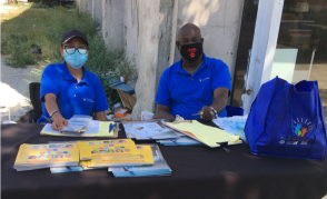 Two people wearing matching shirts and face masks sit at a community outreach event table