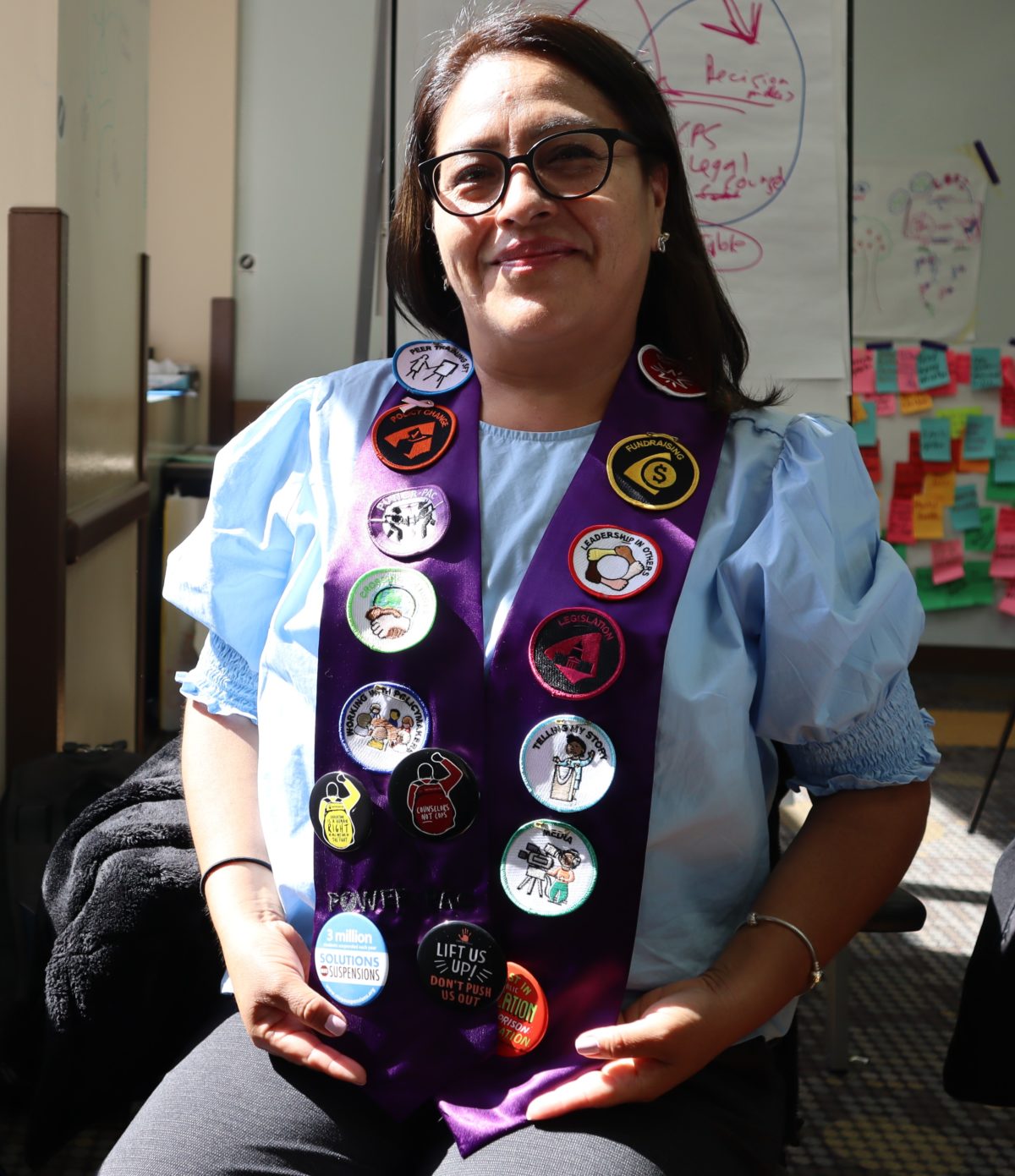 Una mujer latina muestra con orgullo su faja morada cubierta con insignias de líder avanzado