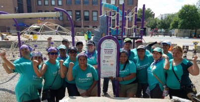 A group of about 15 volunteers, clad in matching teal shirts, pose in front of a playground they are building. Some have their arms flexed to signify strength.