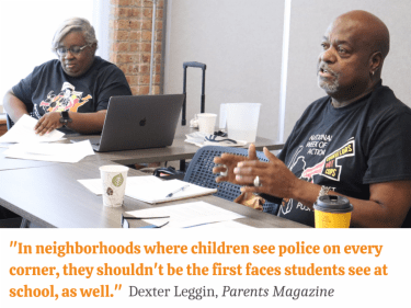 A woman and man, sitting at a table, present during a meeting. Below the image is a quote by Dexter Leggin: In neighborhoods where children see police on every corner, they shouldn't be the first faces students see at school, as well.
