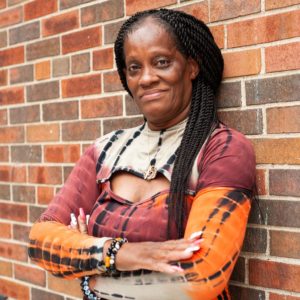 A woman leans against a brick wall with her arms crossed