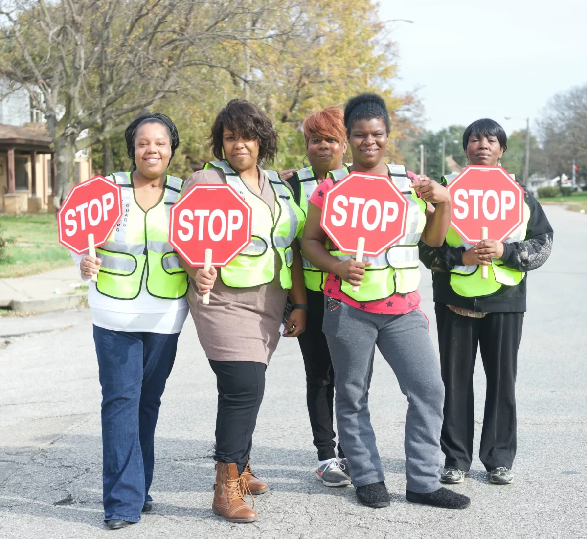 Five women wearing safety vests and holding paper stop signs stand in the middle of the street