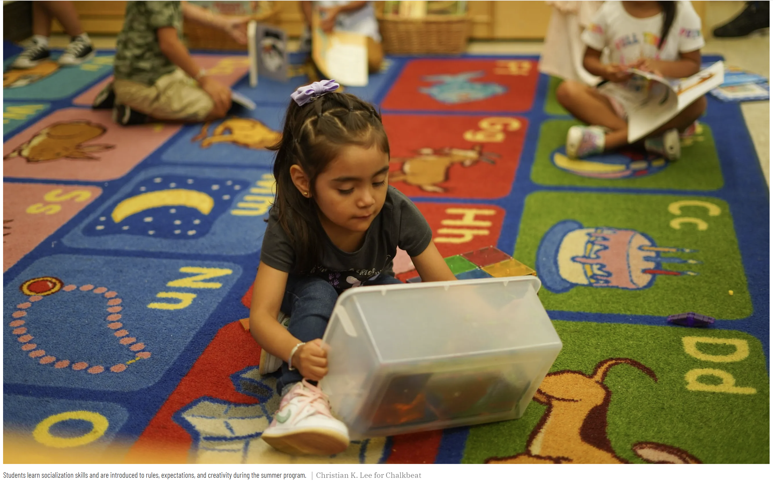 A young girl sits on a colorful rug in preschool and looks through a plastic bin filled with toys and other learning tools. Photo credit: Chalkbeat
