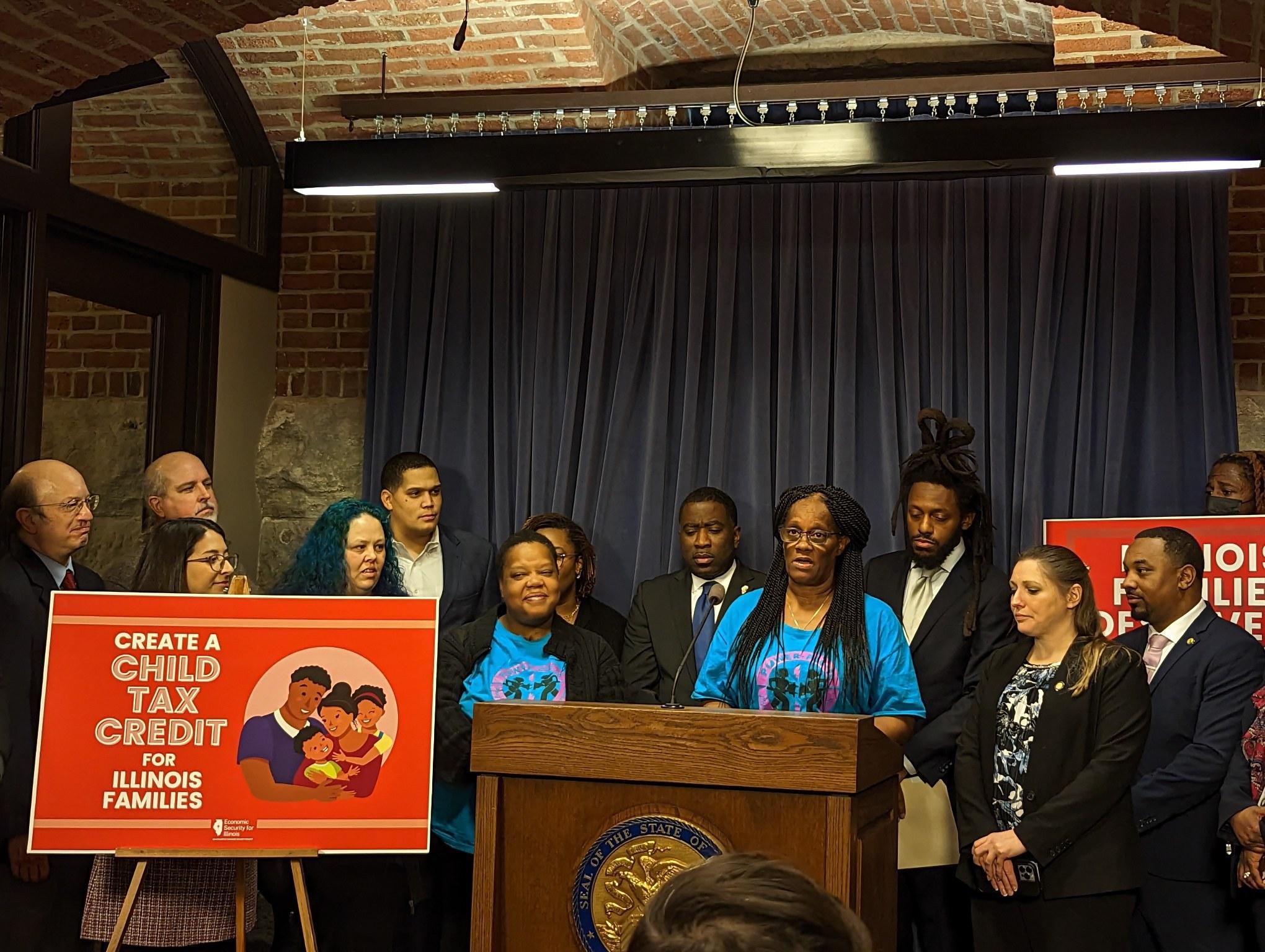 Una mujer afroamericana, líder de padres de POWER-PAC IL, habla en apoyo del Crédito Tributario por Hijos en un podio en el Capitolio del Estado de Illinois. De pie a su alrededor hay otros miembros de POWER-PAC IL, personal de COFI, legisladores estatales y defensores de la comunidad.