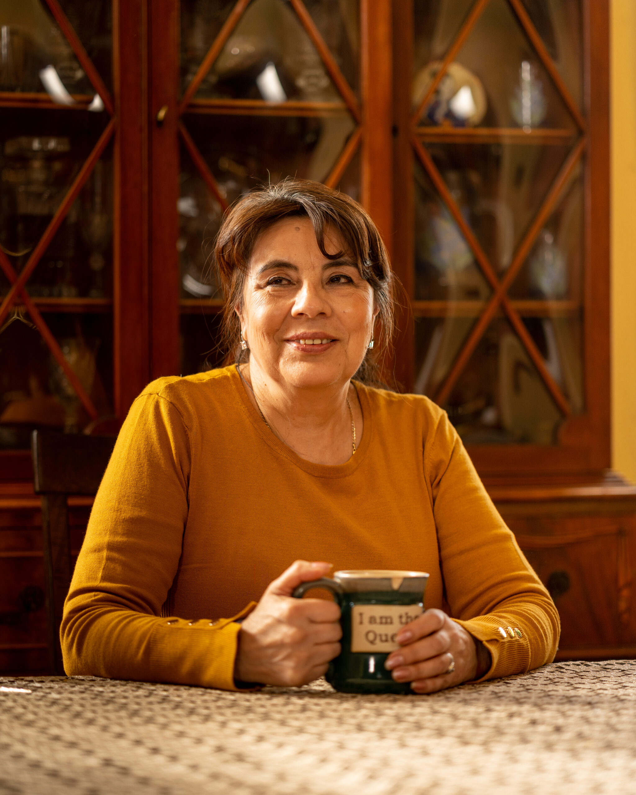 Una mujer latina con un suéter amarillo sostiene una taza de café y se sienta en una mesa