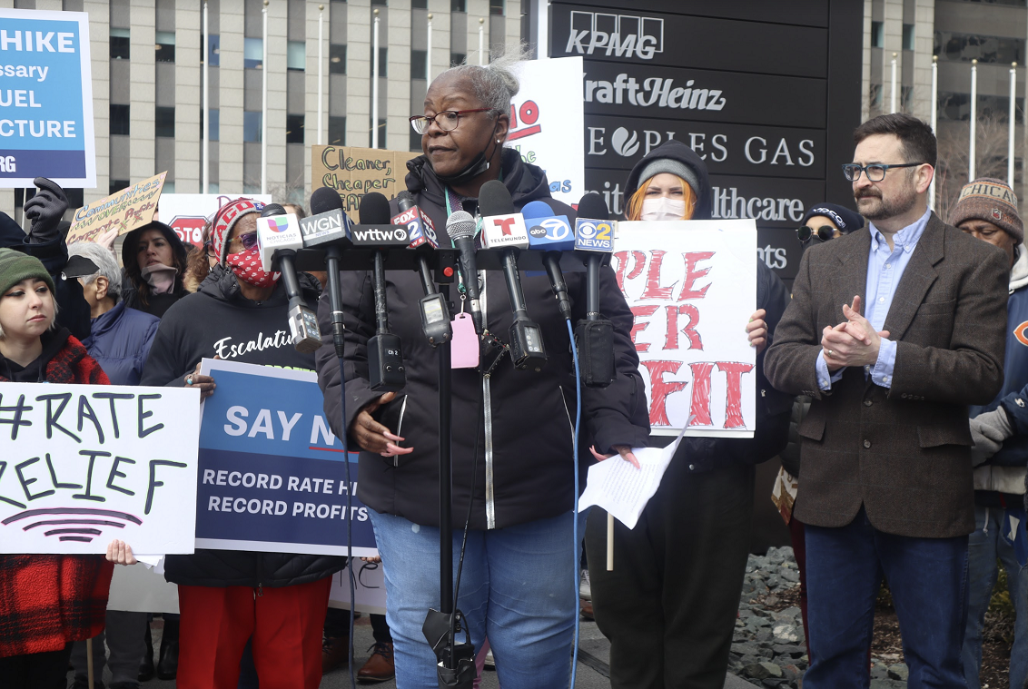 An African-American woman stands at a podium behind various news station microphones and speaks about her bad experiences with Peoples Gas. She is surrounded by fellow community advocates.