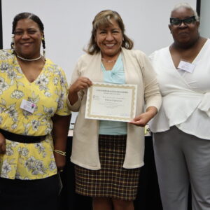 Three women pose for a photo at the end of Phase 3 training for The COFI Way. The woman in the middle holds up a certificate of completion.