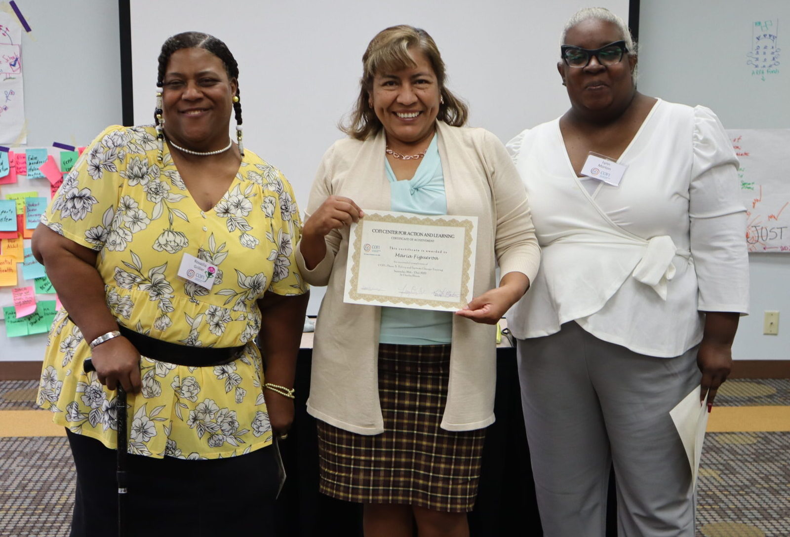 Three women pose for a photo at the end of Phase 3 training for The COFI Way. The woman in the middle holds up a certificate of completion.