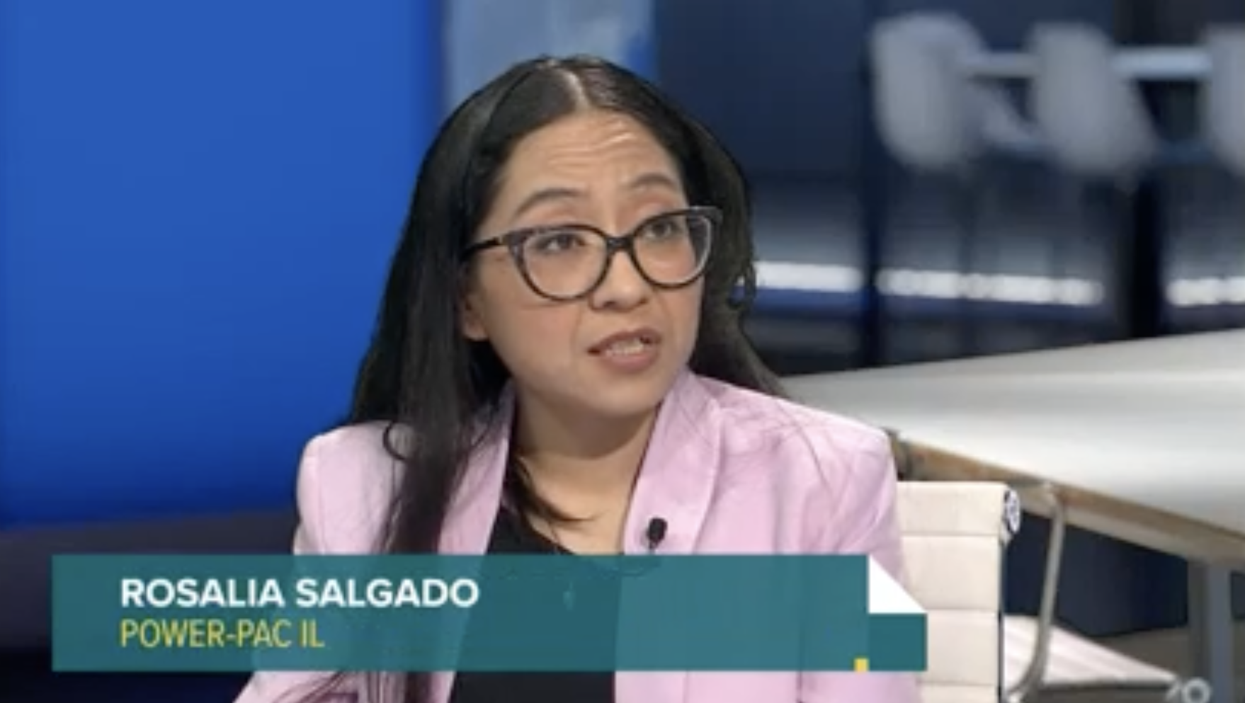 Una mujer latina con un blazer rosa y anteojos es entrevistada en una estación de noticias de la televisión pública local.