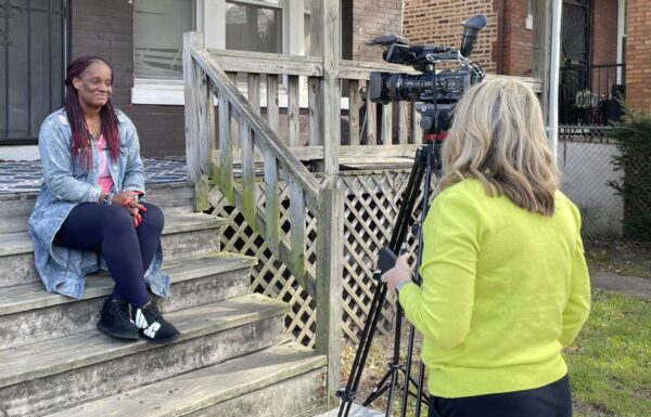 Donna Carpenter sits on the steps outside her home and is speaking to a journalist. There is a TV camera pointed at Donna.