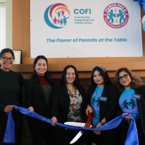 Illinois State Senator Karina Villa and Illinois State Representative Barbara Hernandez join COFI parents Nelly and Liliana for a ribbon cutting. The women are holding a blue ribbon and scissors.