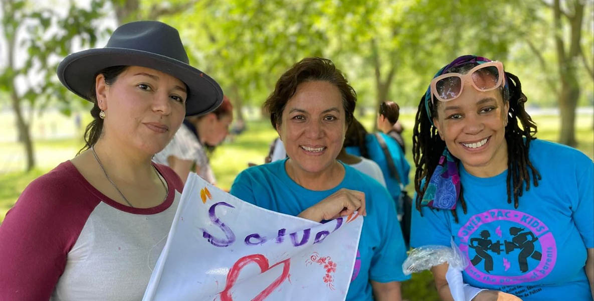 Three women smile and pose for a group picture. One is holding a flag.