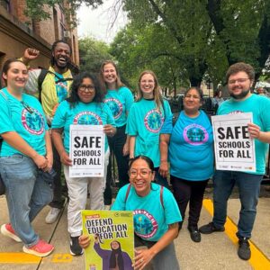 8 people including one Black man, 4 Latina women, and 2 white women, and 1 white male. Some are holding signs that read "Safe Schools for All" and "Defend Education for All"
